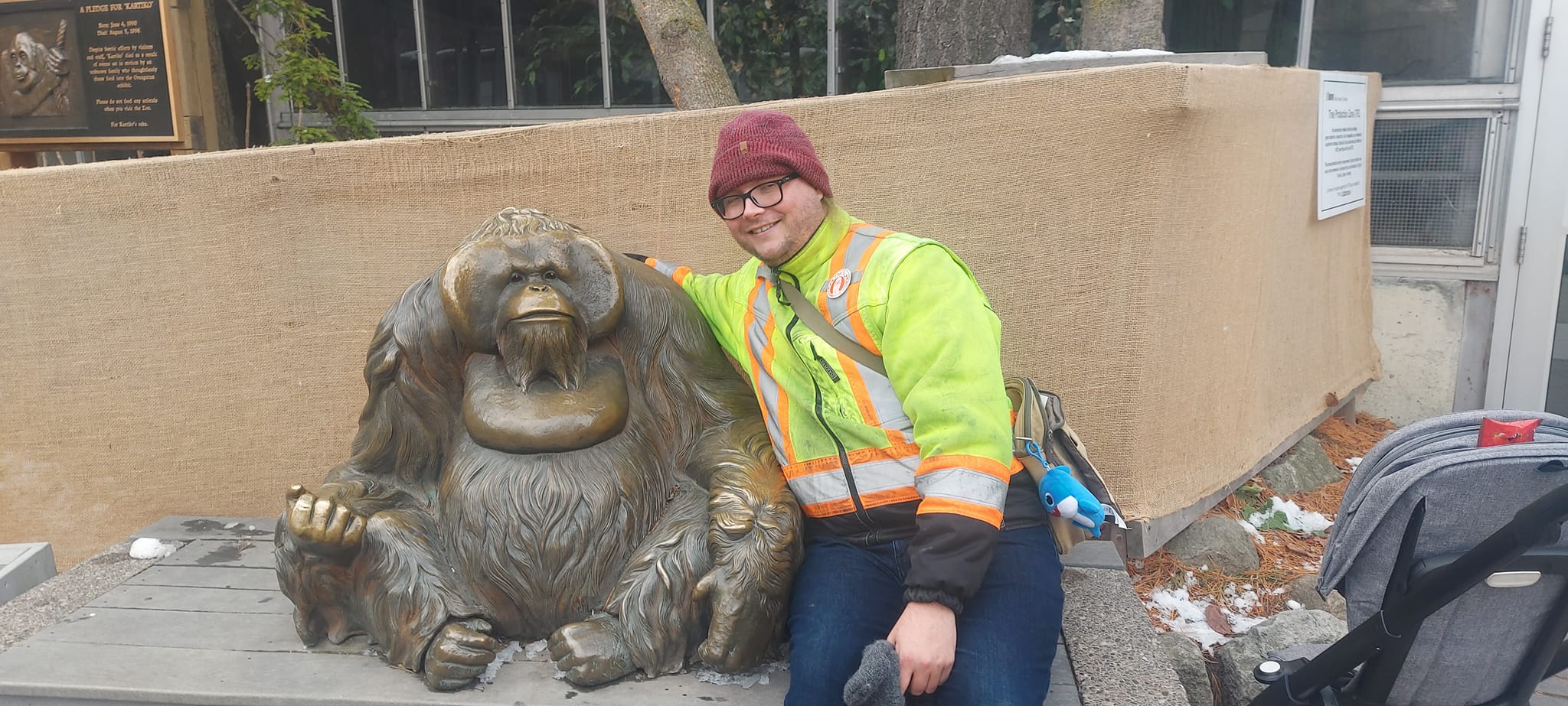 A man wearing a red toque and a high visibility winter coat sits next to an orangutan statue, his arm over its sholder